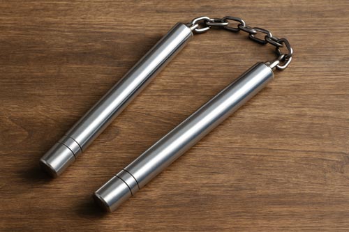 A pair of polished metal nunchucks with a heavy chain connector resting on a wooden surface.
